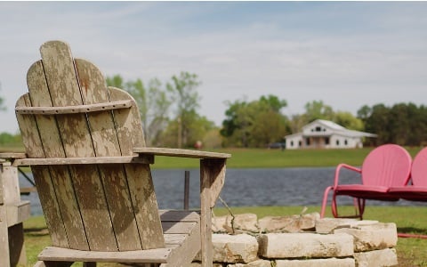 Chairs set up around a fire pit during the day, with a lake and a new rural home in the distance.