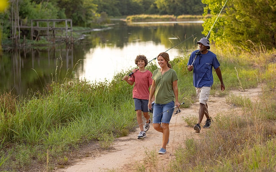 A family of three walks alongside a pond after fishing on their rural property.