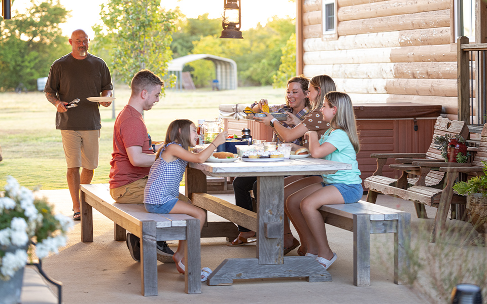 A family of six having dinner at a table outside of their rural home.