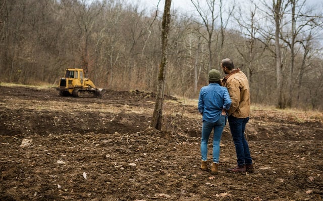 A couple standing outdoors, watching at a bulldozer clear land for their new rural home.
