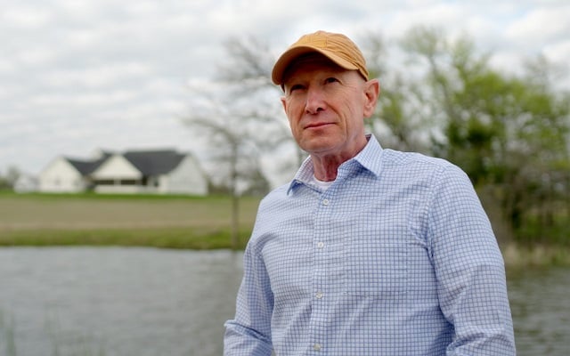 A man in a baseball hat and button down shirt standing in front of a lake with his new rural home in the distance.